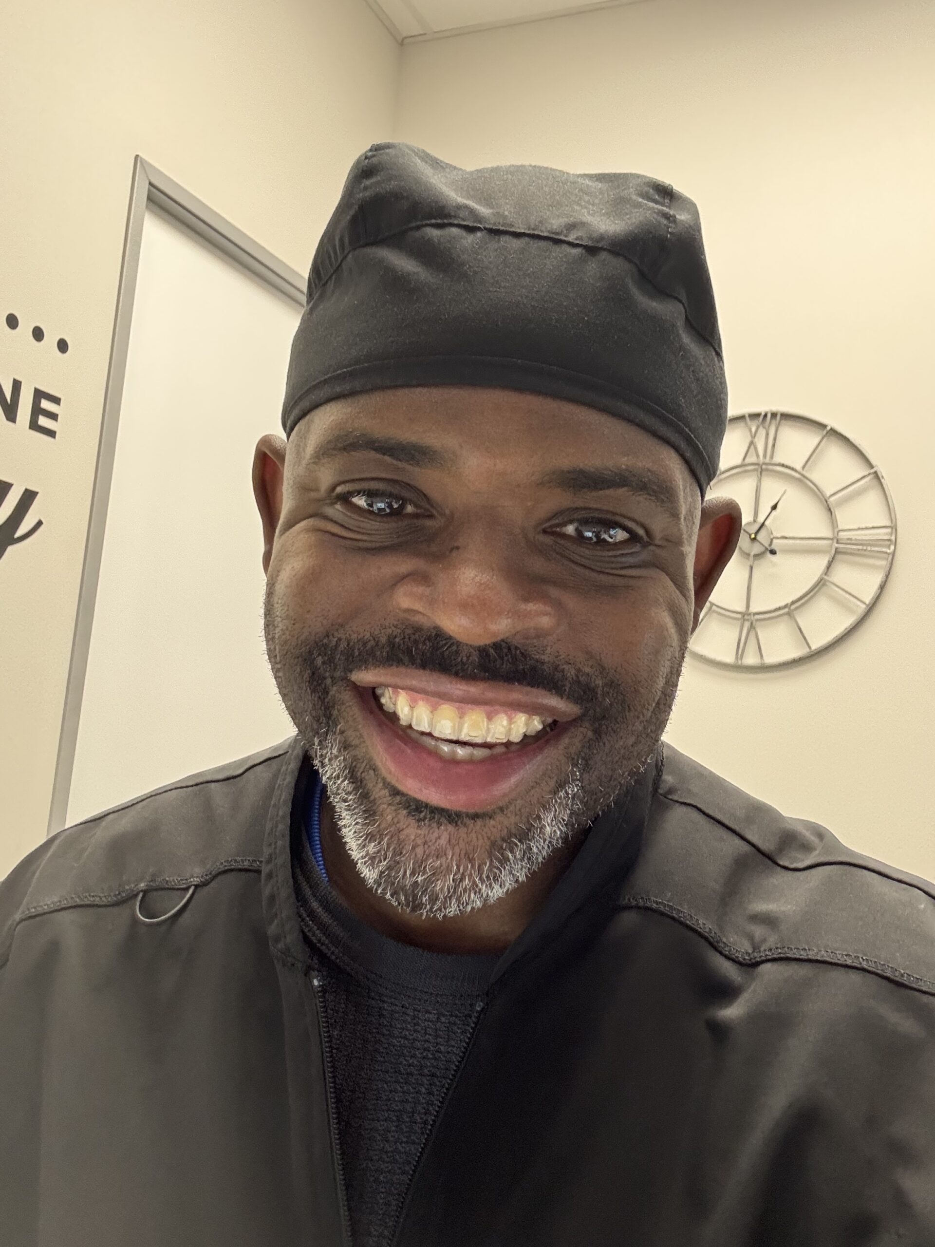 Smiling person in a black chef hat and jacket, indoors, with a clock on the wall behind.
